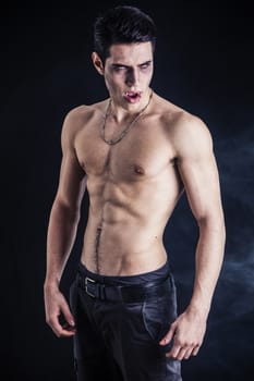 Portrait of a Young Vampire Man Shirtless, Showing his Torso, Chest and Abs, Looking at the Camera, on Dark Background.