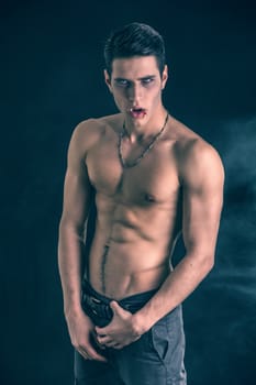 Portrait of a Young Vampire Man Shirtless, Showing his Torso, Chest and Abs, Looking at the Camera, on Dark Background.
