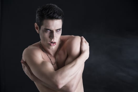 Portrait of a Young Vampire Man Shirtless, Showing his Torso, Chest and Abs, Looking at the Camera, on Dark Background.