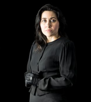 Brunette woman in black standing in front of black backdrop, with her hands held together and looking at the camera with a serious expression on her face.