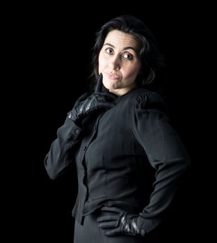 Brunette woman in black standing in front of a black backdrop, one hand on her hip and a finger to her chin with a playful expression on her face.