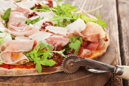 Culinary pizza with prosciutto, dry tomatoes and fresh herbs on wooden cutting board on wooden table. 