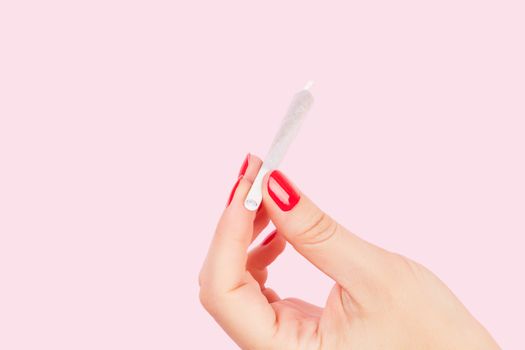 Female hand with red fingernails holding cannabis joint isolated on pink background. Sexy weed smoking.