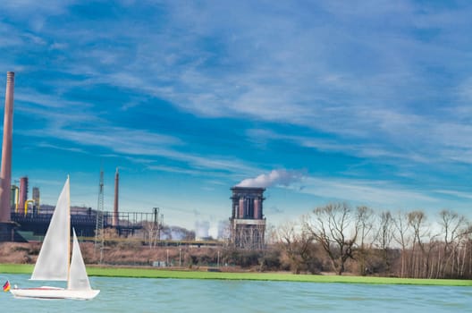 Old power plant on a river against a blue sky. On the water a sail boat. Industry, nature and leisure in harmony.