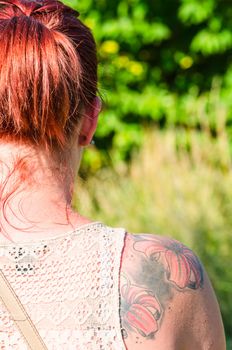 Rotharrige young woman with flowers tattoo on her right shoulder blade. In the background, meadow and trees.