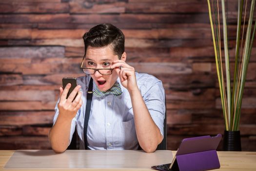 Surprised dapper businesswoman looking over eyeglasses at phone