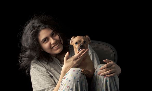 Low key shot of latina teenage girl holding her chihuahua dog and looking at the camera with a smile on her face; the dog's ears are curled back to create a funny appearance and the dog is licking her hand.