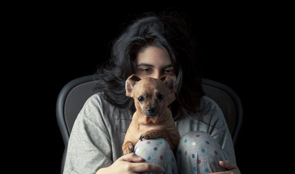Low key shot of latina teenage girl looking at her chihuahua, with her face partially hidden by the dog's head; the dog's ears are curled back to create a funny appearance.