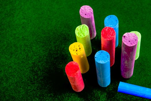 Ten pieces of broken chalk on a green surface. Nine of them, all different colours are standing upright, while one blue piece is lying down.