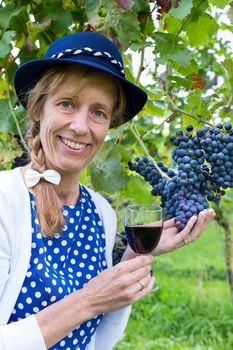 Caucasian middle aged woman holding glass of red wine near bunch of blue grapes in vineyard
