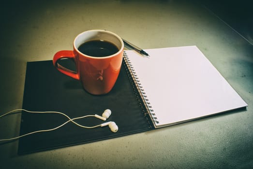 Photograph of an open notebook, a cup of coffee, a pen and a pair of earphones in a grunge style and grainy look