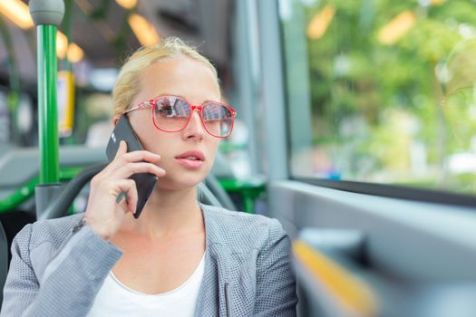 Blonde caucasian business woman talking on cell phone, traveling by bus. Public transport and commuting to work.