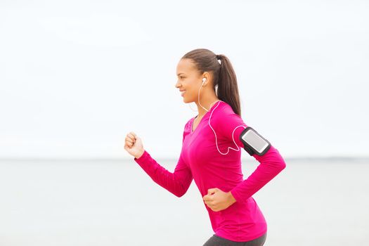 sport, fitness, health, technology and people concept - smiling young african american woman running with smartphone and earphones outdoors