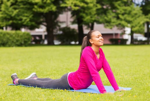 fitness, sport, training, park and lifestyle concept - smiling african american woman stretching on mat outdoors