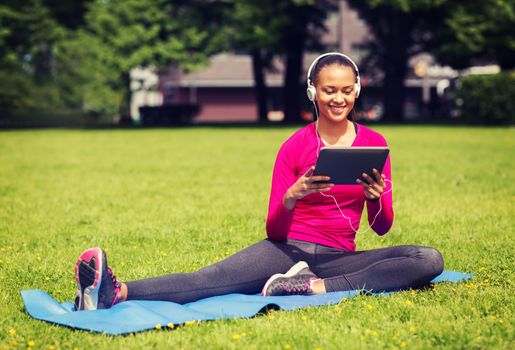 fitness, park, technology and sport concept - smiling african american woman with tablet pc computer and headphones on mat outdoors