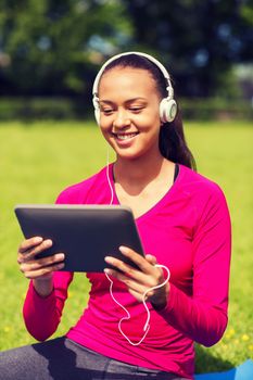 fitness, park, technology and sport concept - smiling african american woman with tablet pc computer and headphones on mat outdoors