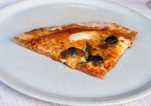 In the picture a slice of pizza with tomato, mozzarella cheese and olives served on a white dish at the restaurant.