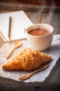 Butter croissant with a cup of hot chocolate and small notebook on wood table with morning scene