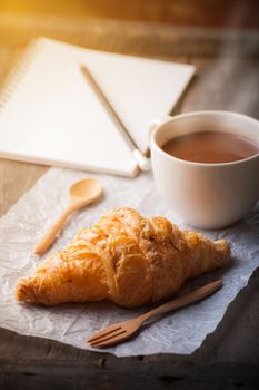 Butter croissant with a cup of hot chocolate and small notebook on wood table with morning scene
