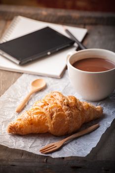 Butter croissant with a cup of hot chocolate and small notebook on wood table with morning scene