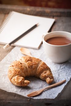 Butter croissant with a cup of hot chocolate and small notebook on wood table with morning scene