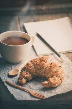 Butter croissant with a cup of hot chocolate and small notebook on wood table in morning time with film filter effect
