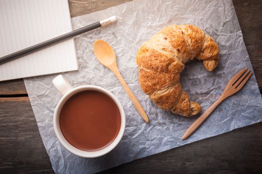 Butter croissant with a cup of hot chocolate and small notebook on wood table with morning scene