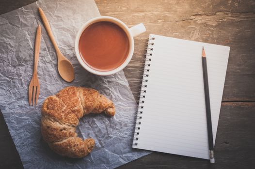 Butter croissant with a cup of hot chocolate and small notebook with blank area for text or message on wood table in morning time with film filter effect