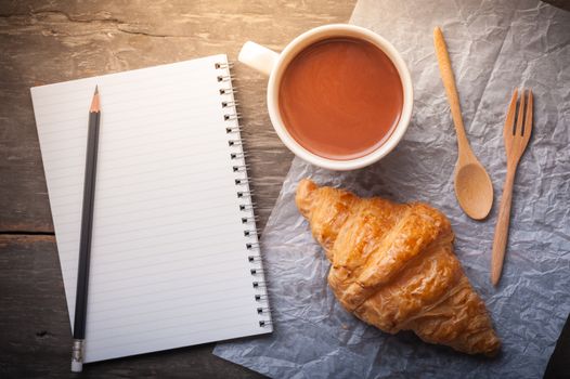 Butter croissant with a cup of hot chocolate and small notebook with blank area for text or message on wood table in morning time