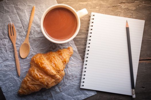 Butter croissant with a cup of hot chocolate and small notebook with blank area for text or message on wood table in morning time
