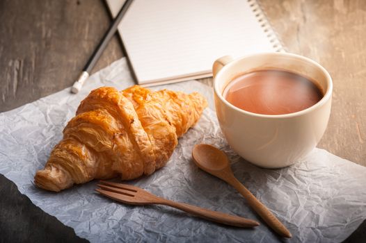 Butter croissant with a cup of hot chocolate and small notebook on wood table with morning scene