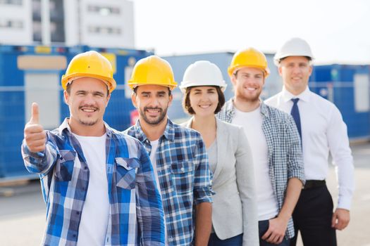 business, building, teamwork, gesture and people concept - group of smiling builders in hardhats showing thumbs up outdoors