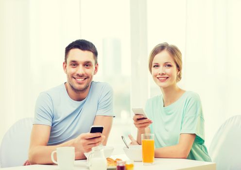 food, home, couple and technology concept - smiling couple with smartphones reading news and having breakfast at home