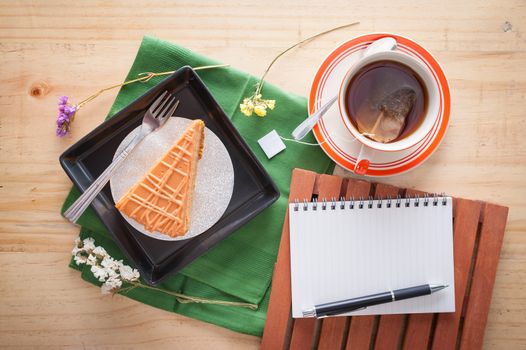 a piece of Thai tea cake on black ceramic dish with a cup of hot tea and notepad on wood table in afternoon