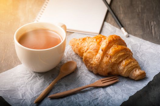 Butter croissant with a cup of hot chocolate and small notebook on wood table with morning scene