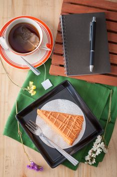 a piece of Thai tea cake on black ceramic dish with a cup of hot tea and notepad on wood table in afternoon