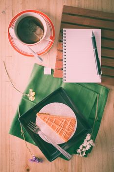 a piece of Thai tea cake on black ceramic dish with a cup of hot tea and notepad on wood table in afternoon with film filter effect