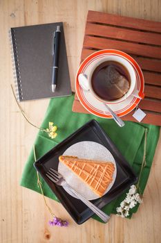 a piece of Thai tea cake on black ceramic dish with a cup of hot tea and notepad on wood table in afternoon