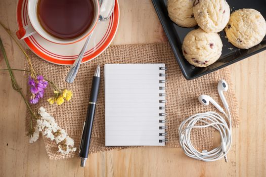 Opened notepad with pen, cookies in black ceramic dish, small earphone, and a cup of hot tea on wood table in cafe with morning scene