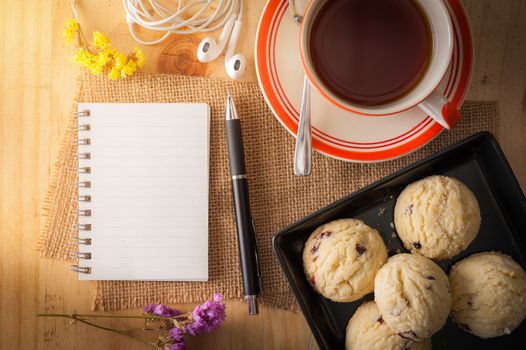 Opened notepad with pen, cookies in black ceramic dish, small earphone, and a cup of hot tea on wood table in cafe with morning scene