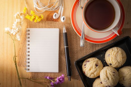 Opened notepad with pen, cookies in black ceramic dish, small earphone, and a cup of hot tea on wood table in cafe with morning scene