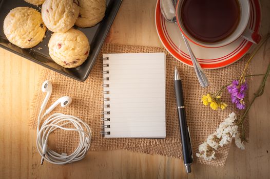 Opened notepad with pen, cookies in black ceramic dish, small earphone, and a cup of hot tea on wood table in cafe with morning light effect