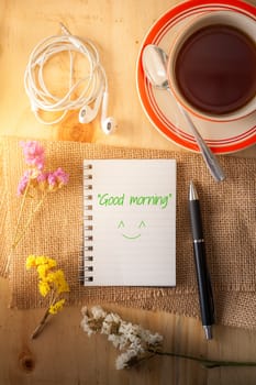 Opened notepad with pen, small earphone, and a cup of hot tea on wood table in cafe with morning scene