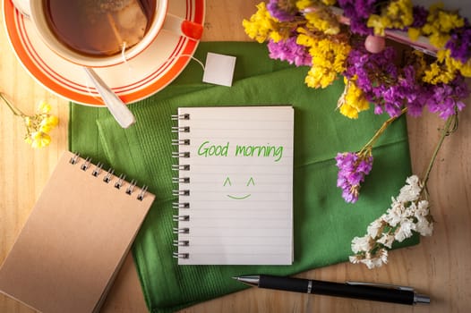"Good morning" is written on opened notepad with pen and a cup of hot tea on wood table in cafe with morning scene
