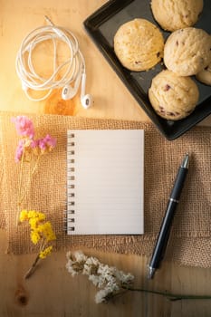 Opened notepad with pen, cookies in black ceramic dish, small earphone on wood table in cafe with morning scene