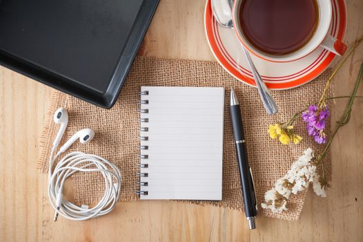 Opened notepad with pen, empty black ceramic dish, small earphone, and a cup of hot tea on wood table in cafe with morning scene
