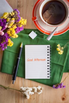 "Good afternoon" is written on opened notepad with pen and a cup of hot tea on wood table in cafe with morning scene