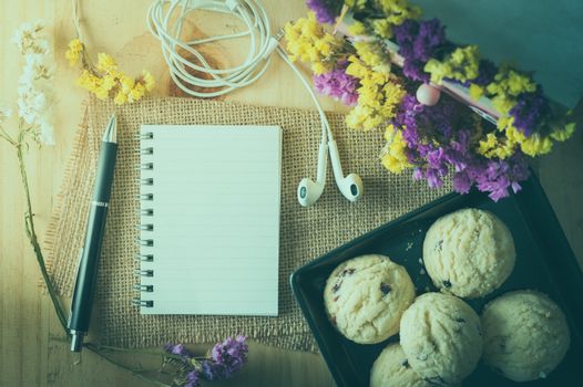 Opened notepad with pen, cookies in black ceramic dish, small earphone, and a cup of hot tea on wood table in cafe with film filter effect
