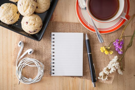 Opened notepad with pen, cookies in black ceramic dish, small earphone, and a cup of hot tea on wood table in cafe with afternoon scene