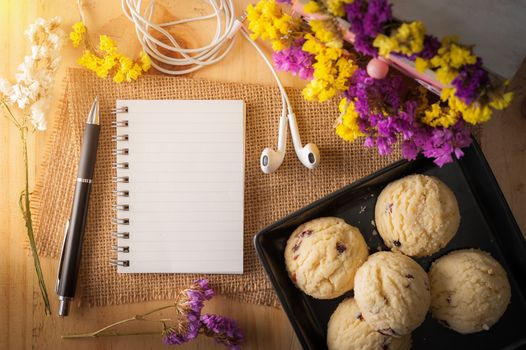 Opened notepad with pen, cookies in black ceramic dish, small earphone, and a cup of hot tea on wood table in cafe with morning scene